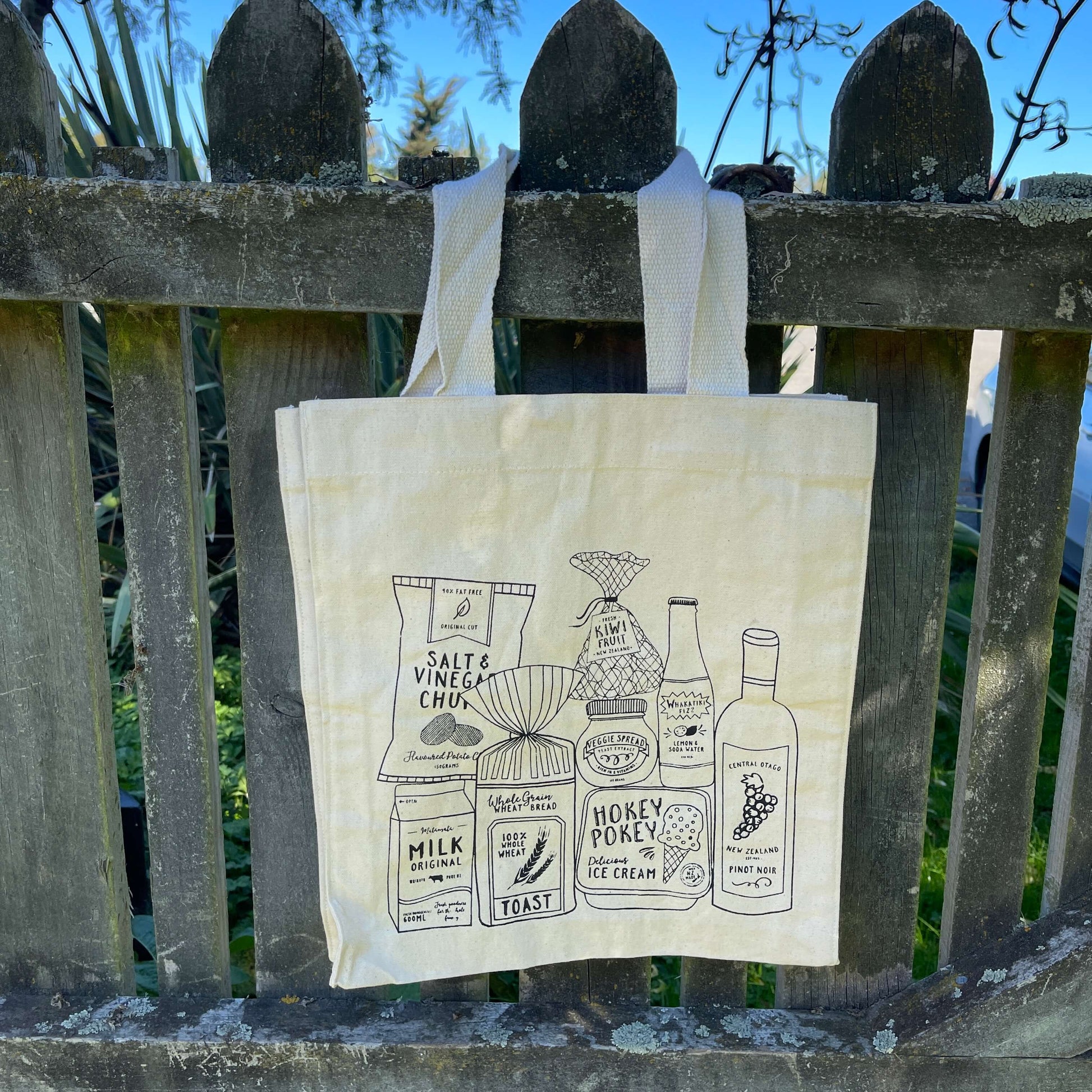 Canvas tote bag with black ink print of various Kiwi grocery staples such as bread, wine & hokey pokey ice cream.