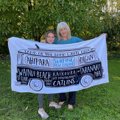 Grandmother & Grandaughter holding a large white beach towel with a Kombi van and names of New Zealand surf locations on it.