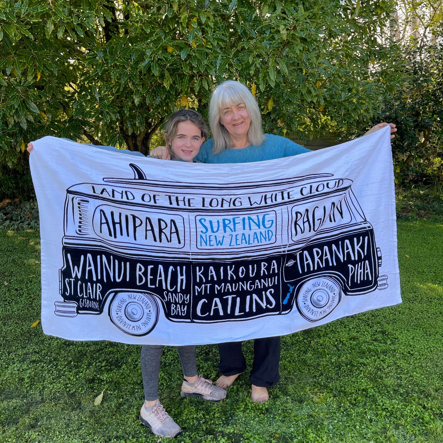 Grandmother & Grandaughter holding a large white beach towel with a Kombi van and names of New Zealand surf locations on it.