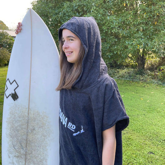 Young girl wearing a black towel hoodie with the words Moana RD on it in white and a map of New Zealand. She is holding a surfboard.