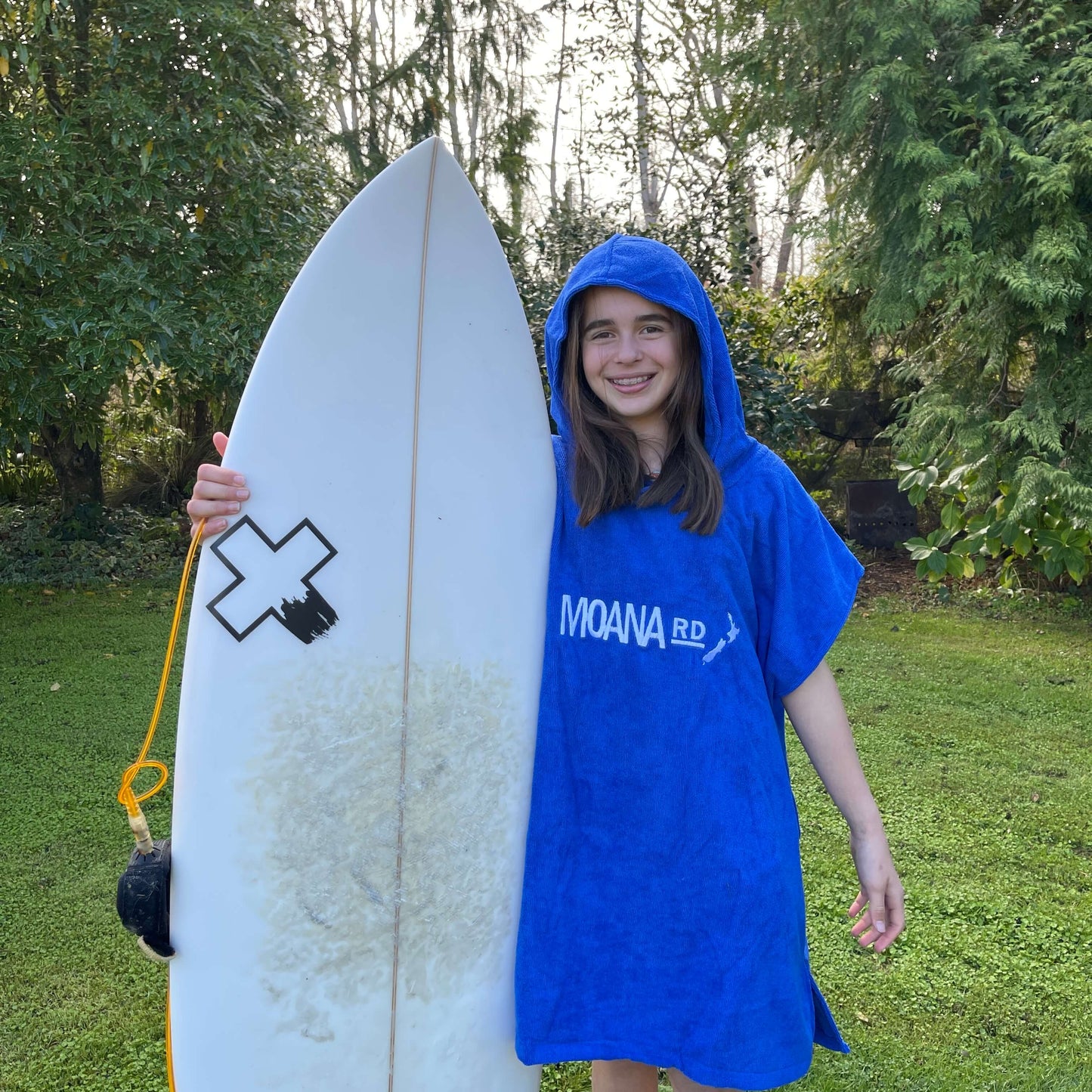 Young girl wearing a blue towel hoodie with the words Moana RD on it in white and a map of New Zealand. She is holding a surfboard.
