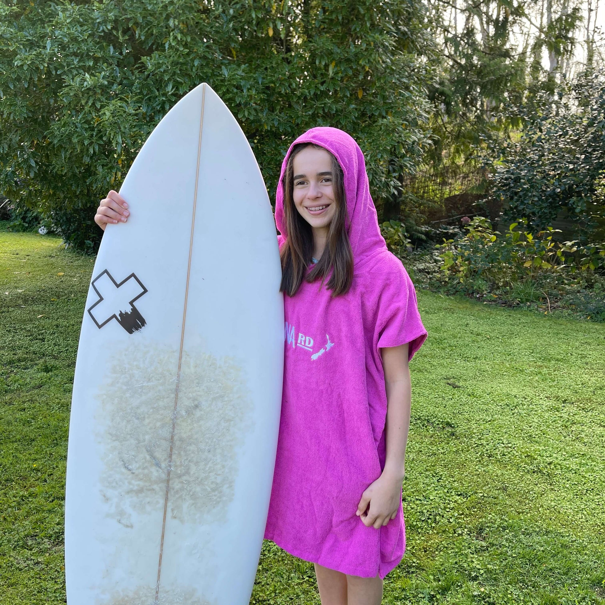 Young girl wearing a bright pink towel hoodie with the words Moana RD on it in white and a map of New Zealand. She is holding a surfboard.