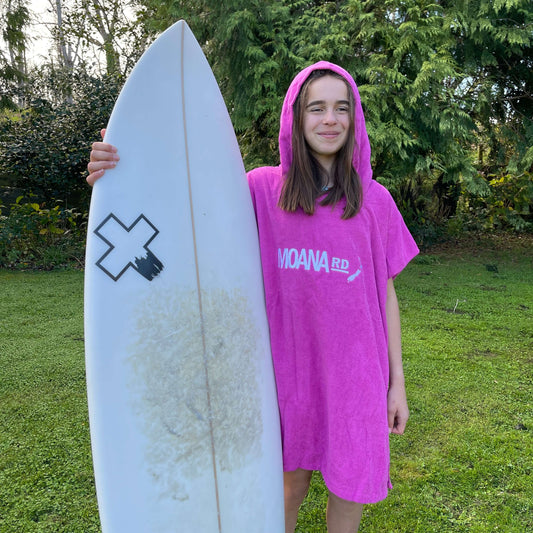 Young girl wearing a bright pink towel hoodie with the words Moana RD on it in white and a map of New Zealand. She is holding a surfboard.