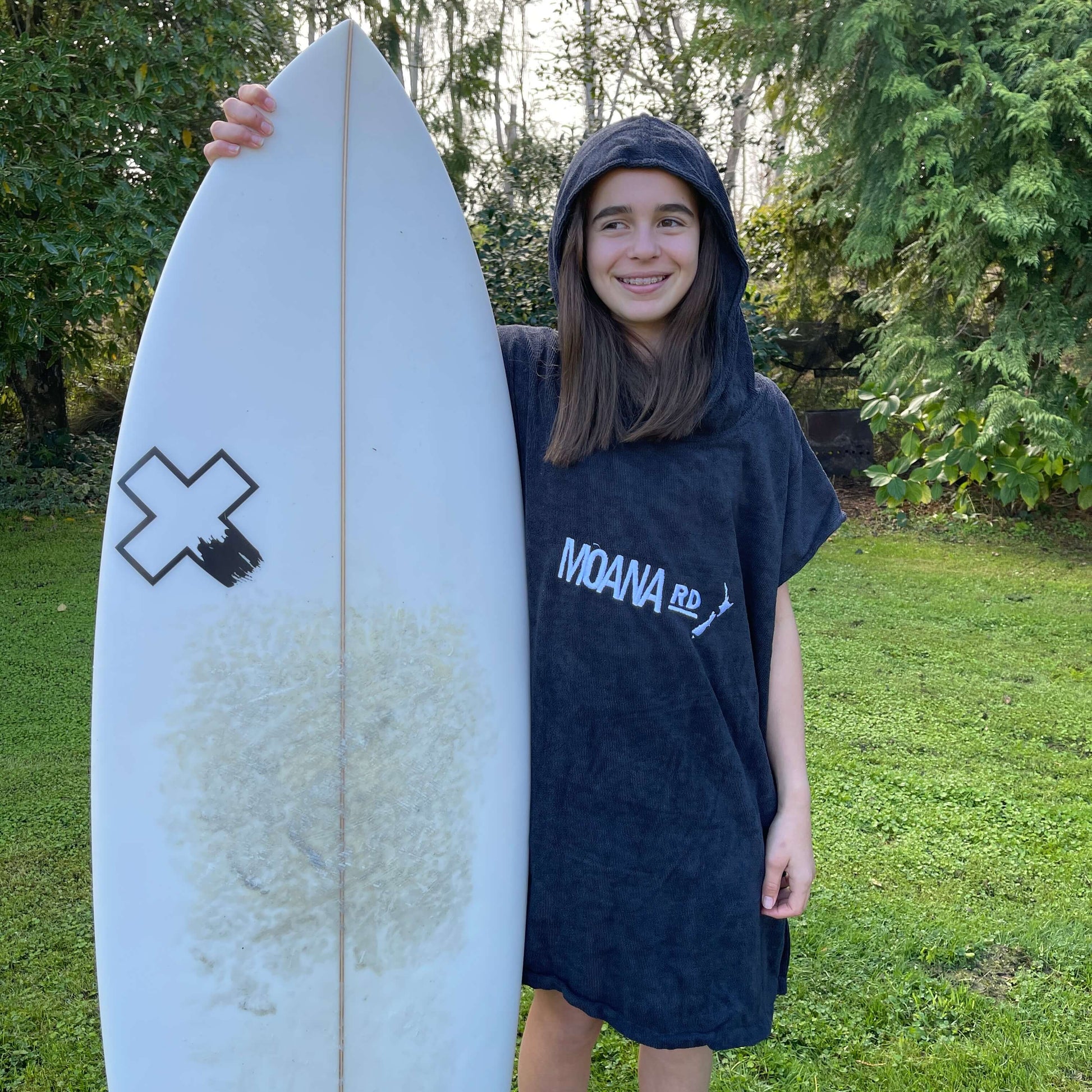 Young girl wearing a black towel hoodie with the words Moana RD on it in white and a map of New Zealand. She is holding a surfboard.