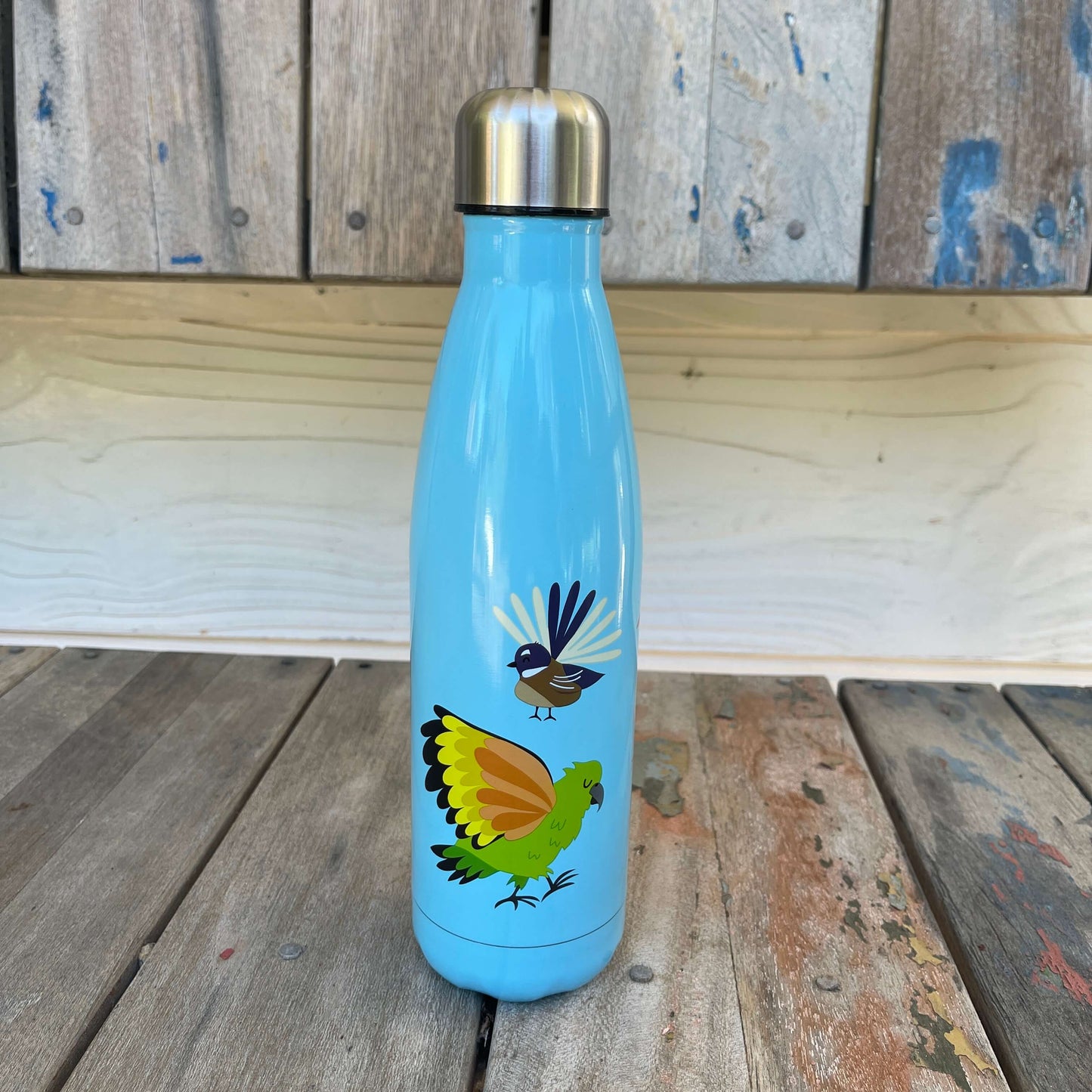 Bright blue drink bottle with a fantail and kea on it sitting on a wooden bench.