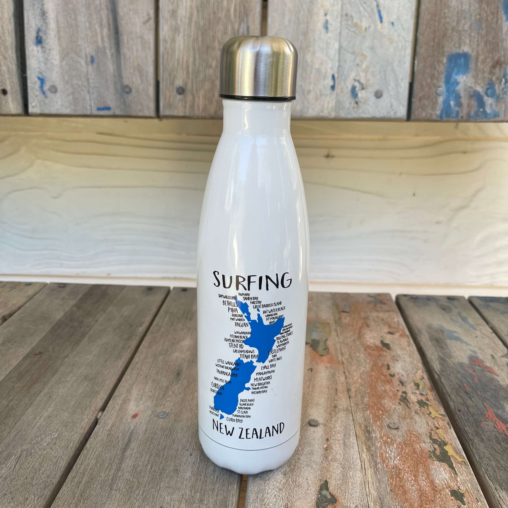 White drink bottle with map of New Zealand featuring surfing hot spots sitting on a wooden bench.