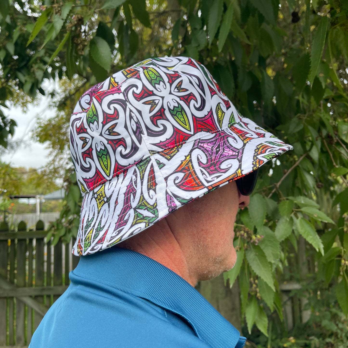 Side view of a mans head wearing a brightly coloured bucket hat with koru patterns.