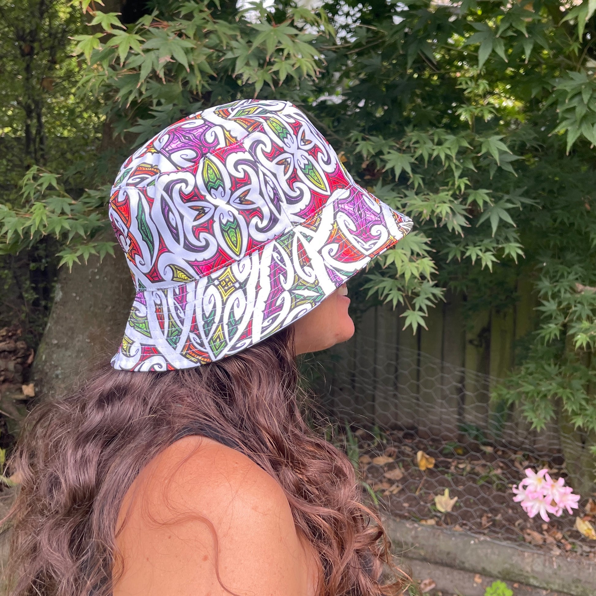 Side profile of a woman with long brown hair head wearing a bucket hat in bright colours with koru patterns in white printed on it.
