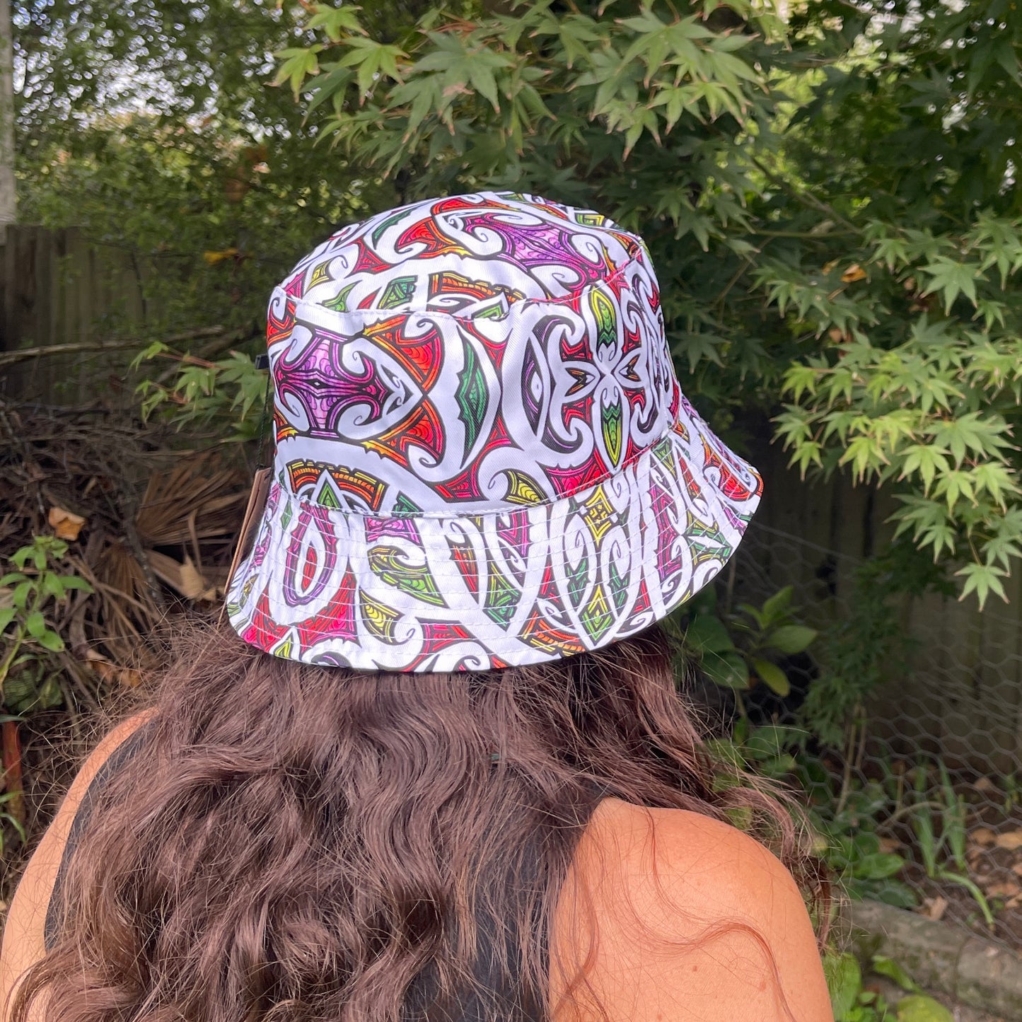 Back view of a woman with long brown hair head wearing a bucket hat in bright colours with koru patterns in white printed on it.