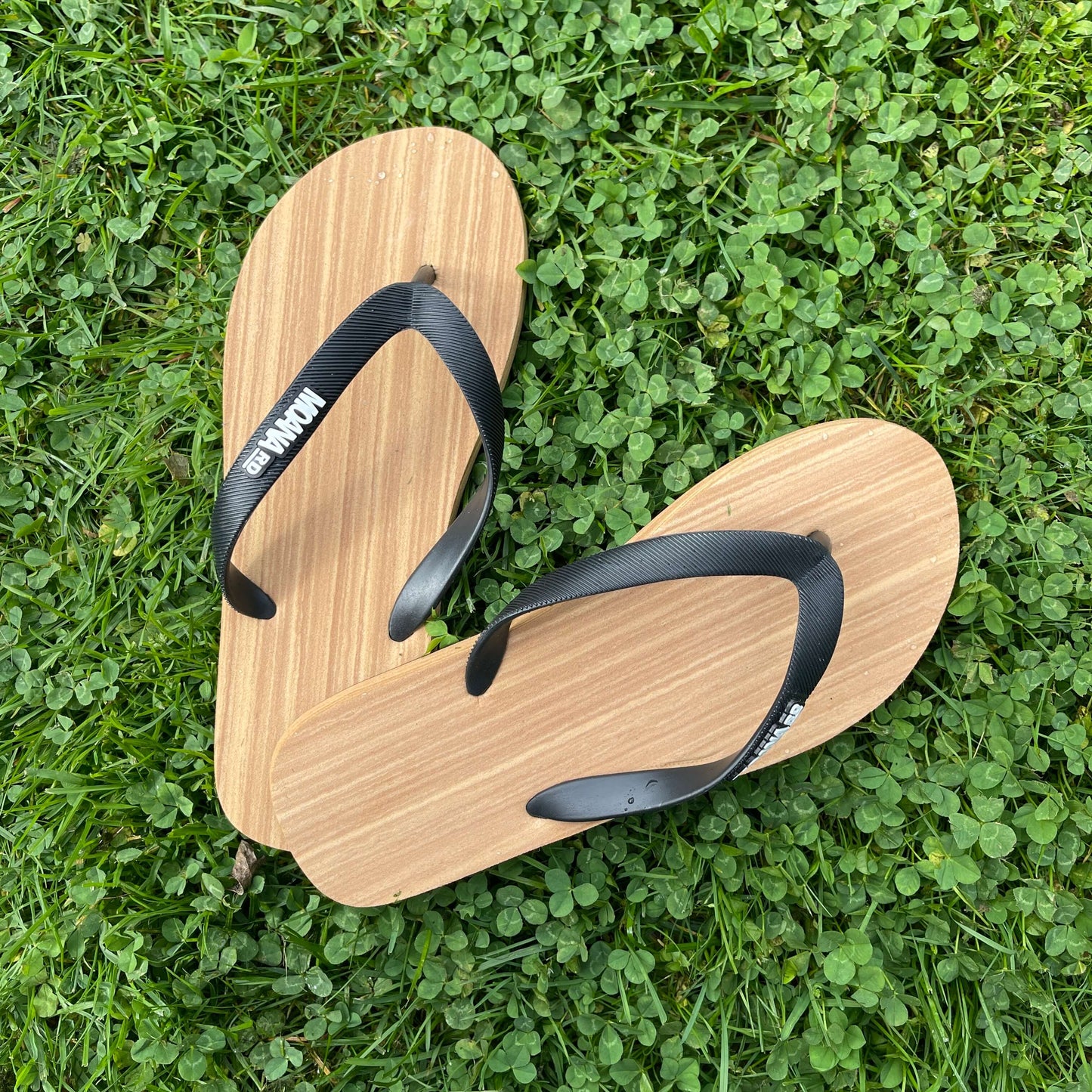 Unisex Jandals with a wood grain look and black straps sitting on grass.