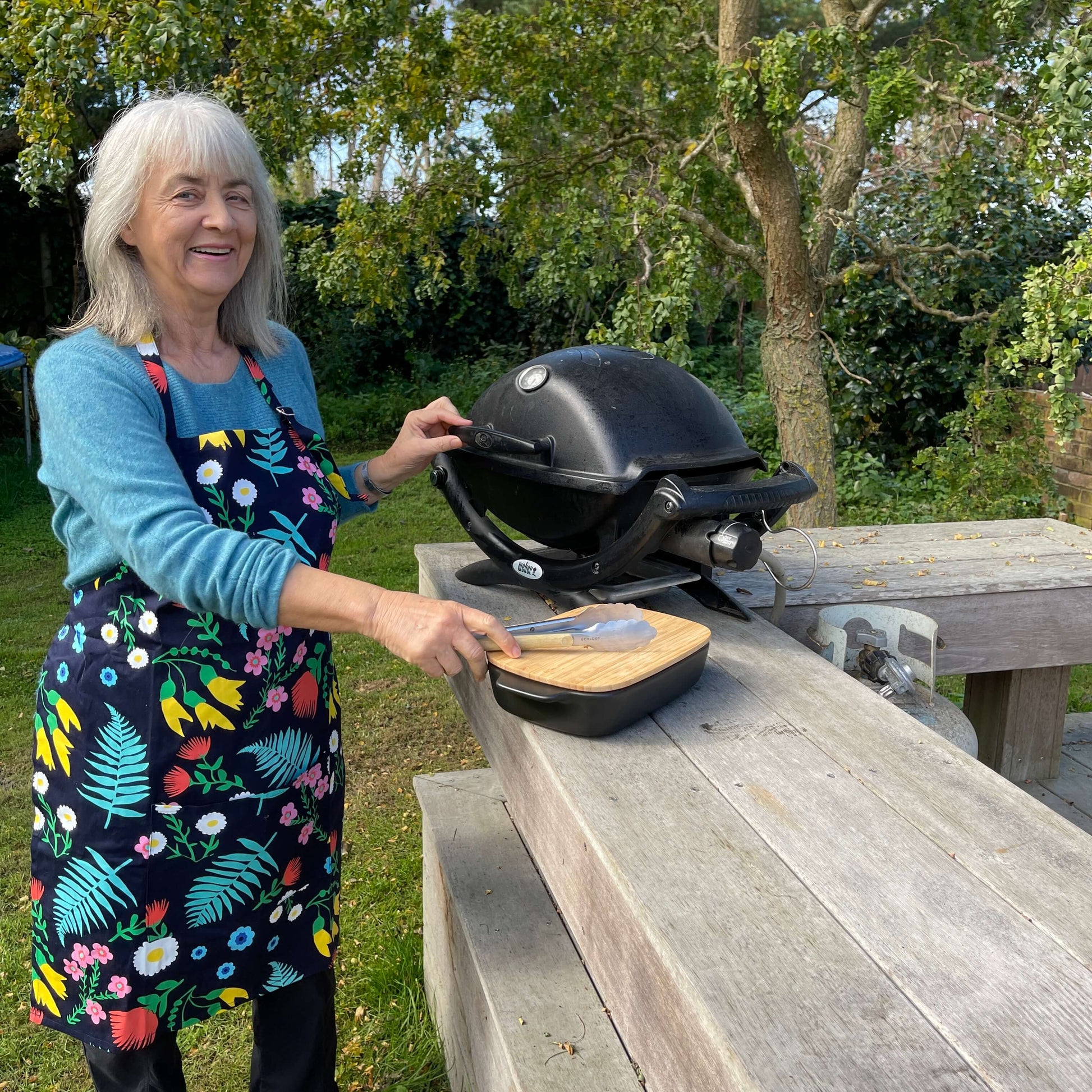 Woman wearing a navy blue and floral apron standing outside at a BBQ.