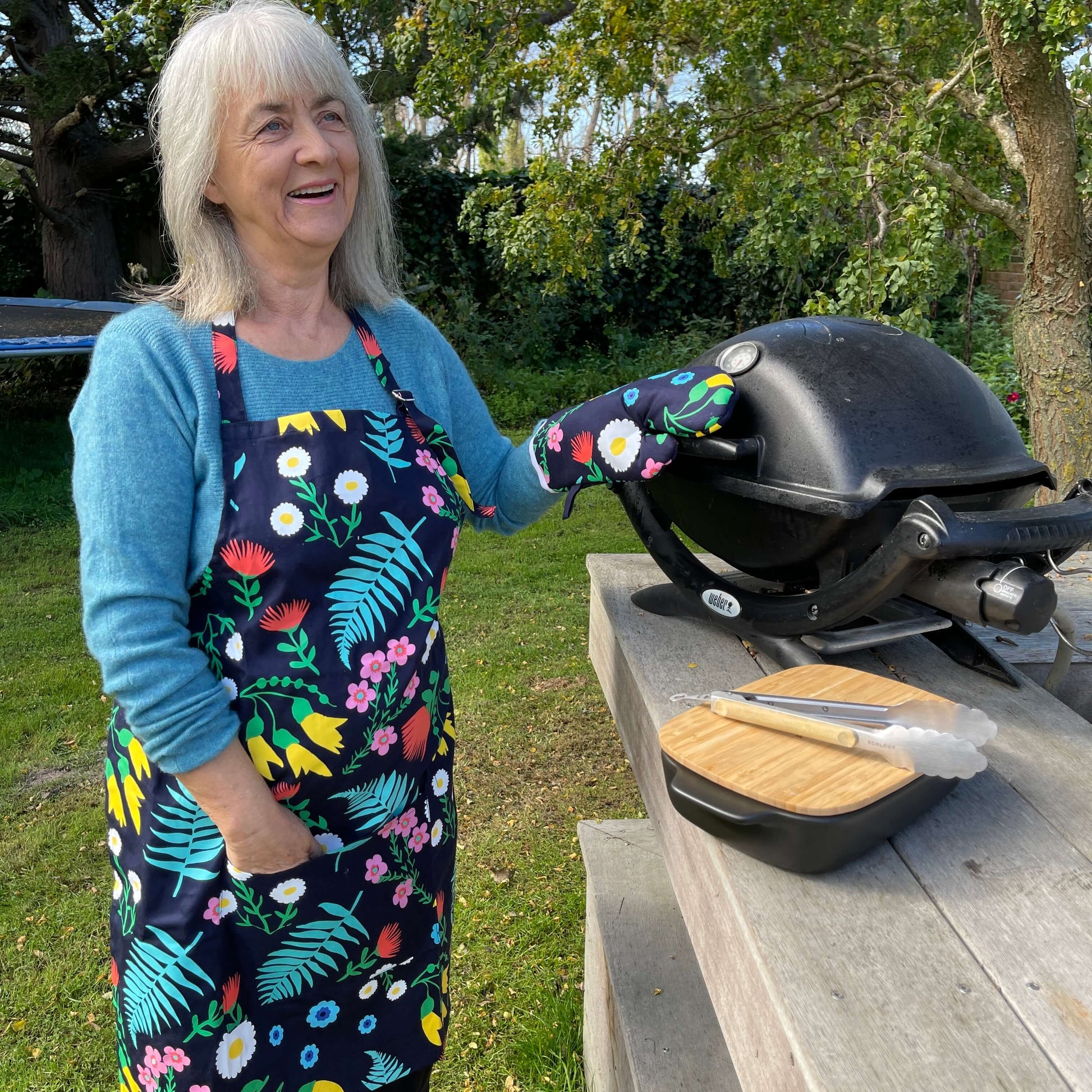 Woman wearing a navy blue and floral apron and mitt set standing outside at a BBQ.