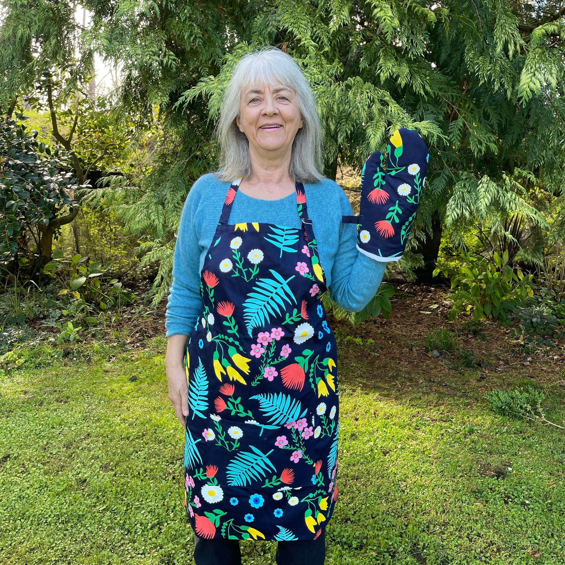 Woman wearing a navy blue and floral apron and mitt set standing outside.