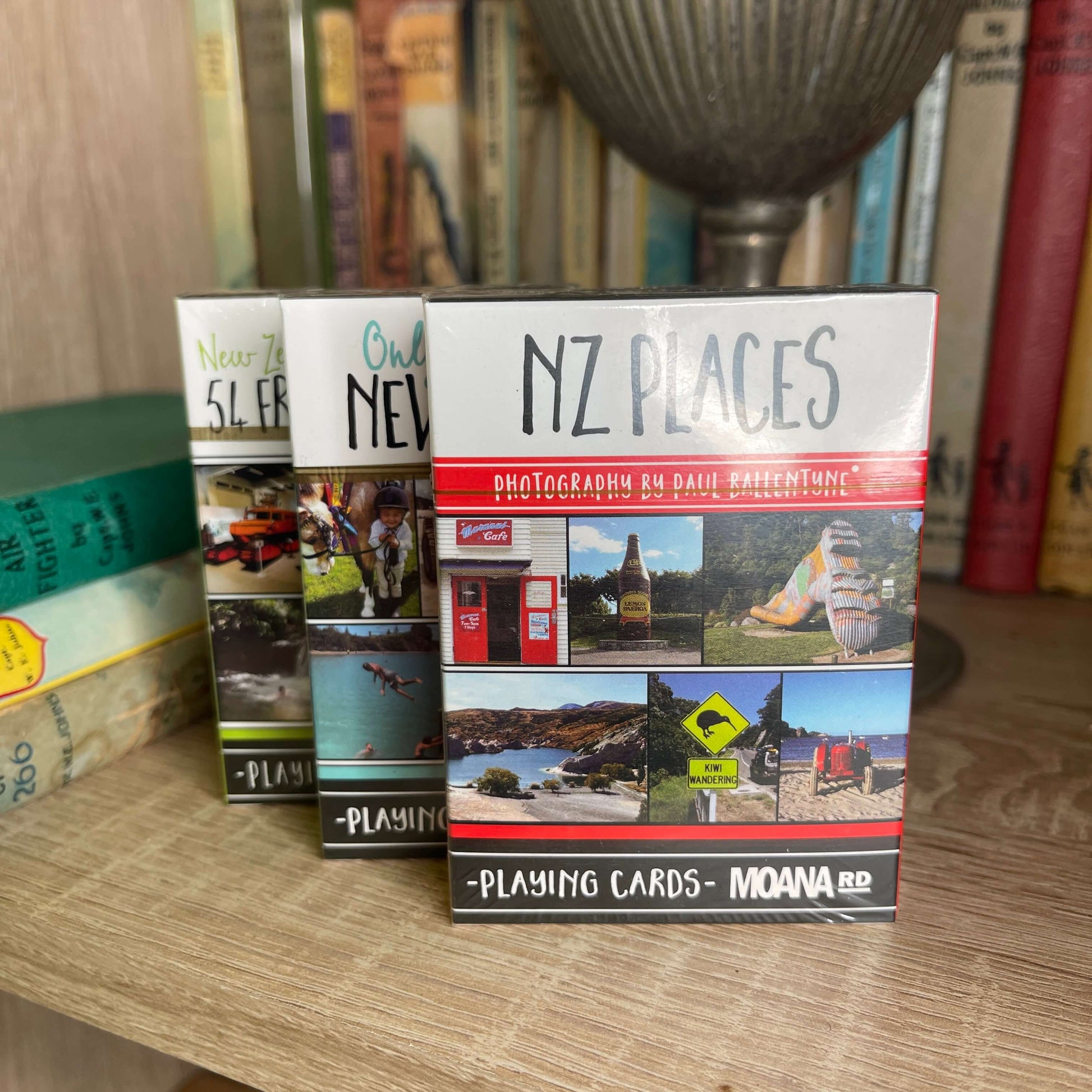 Pack of playing cards featuring NZ places sitting on a book shelf with other card packs behind it.
