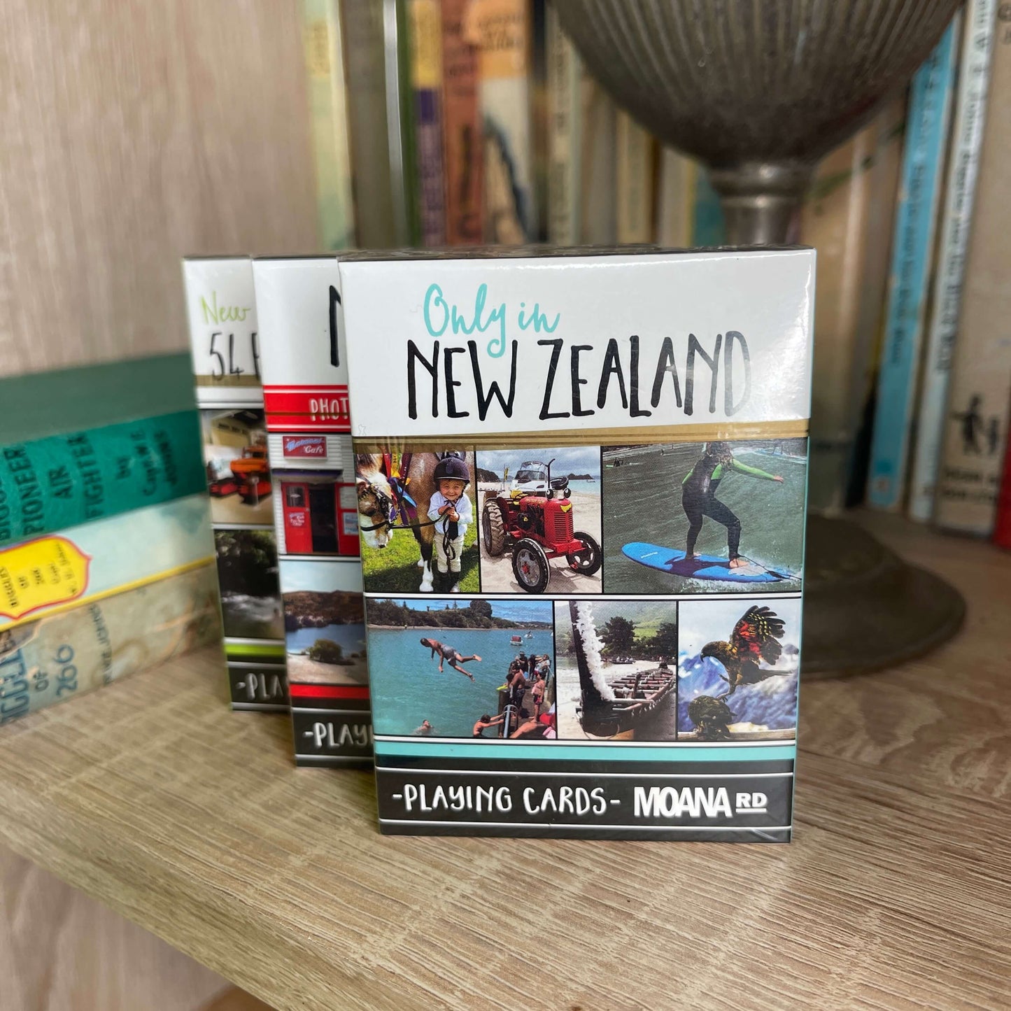 Pack of playing cards featuring scenes only found in New Zealand sitting on a book shelf with other card packs behind it.