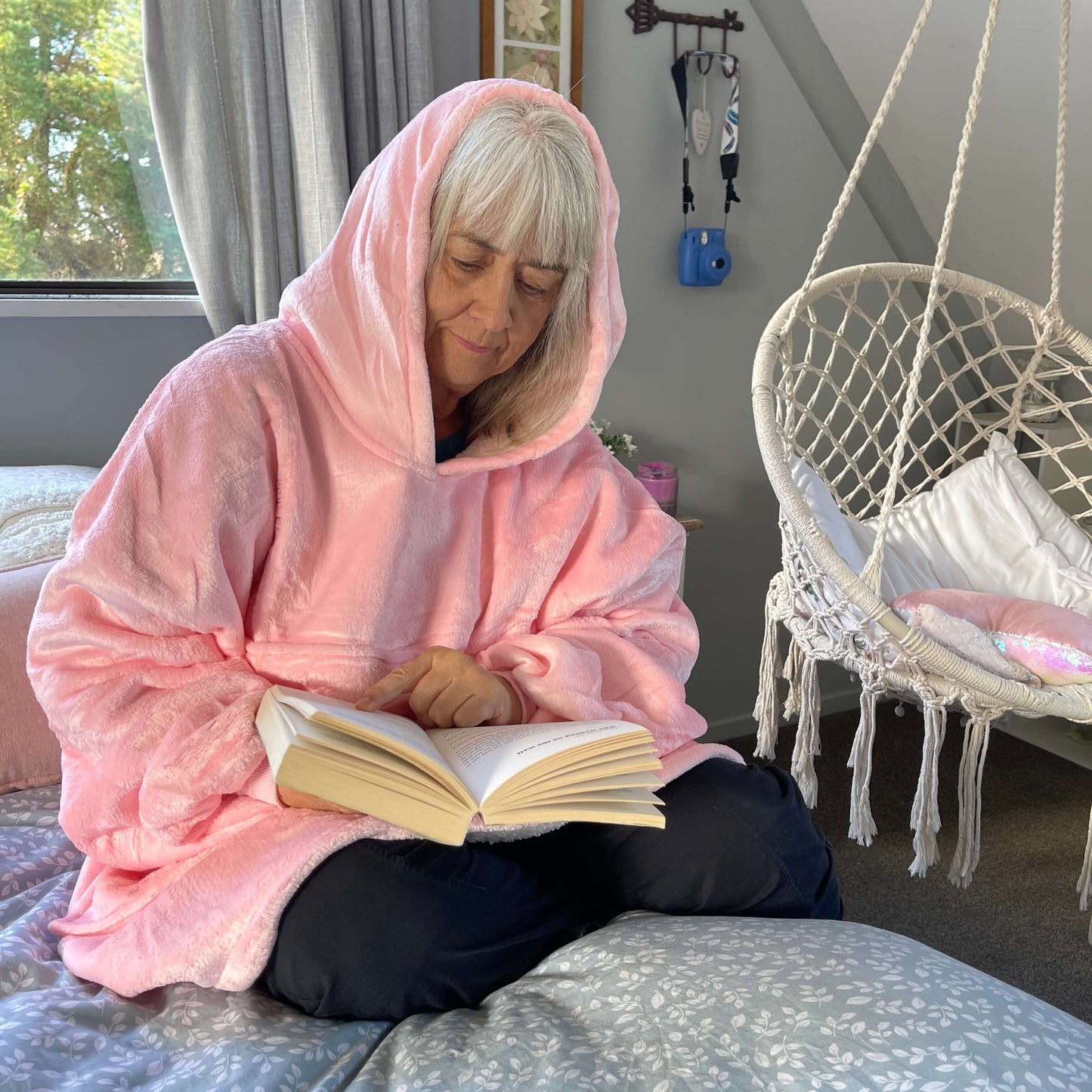 Woman sitting on a bed reading a book wearing an oversized pink hoodie.