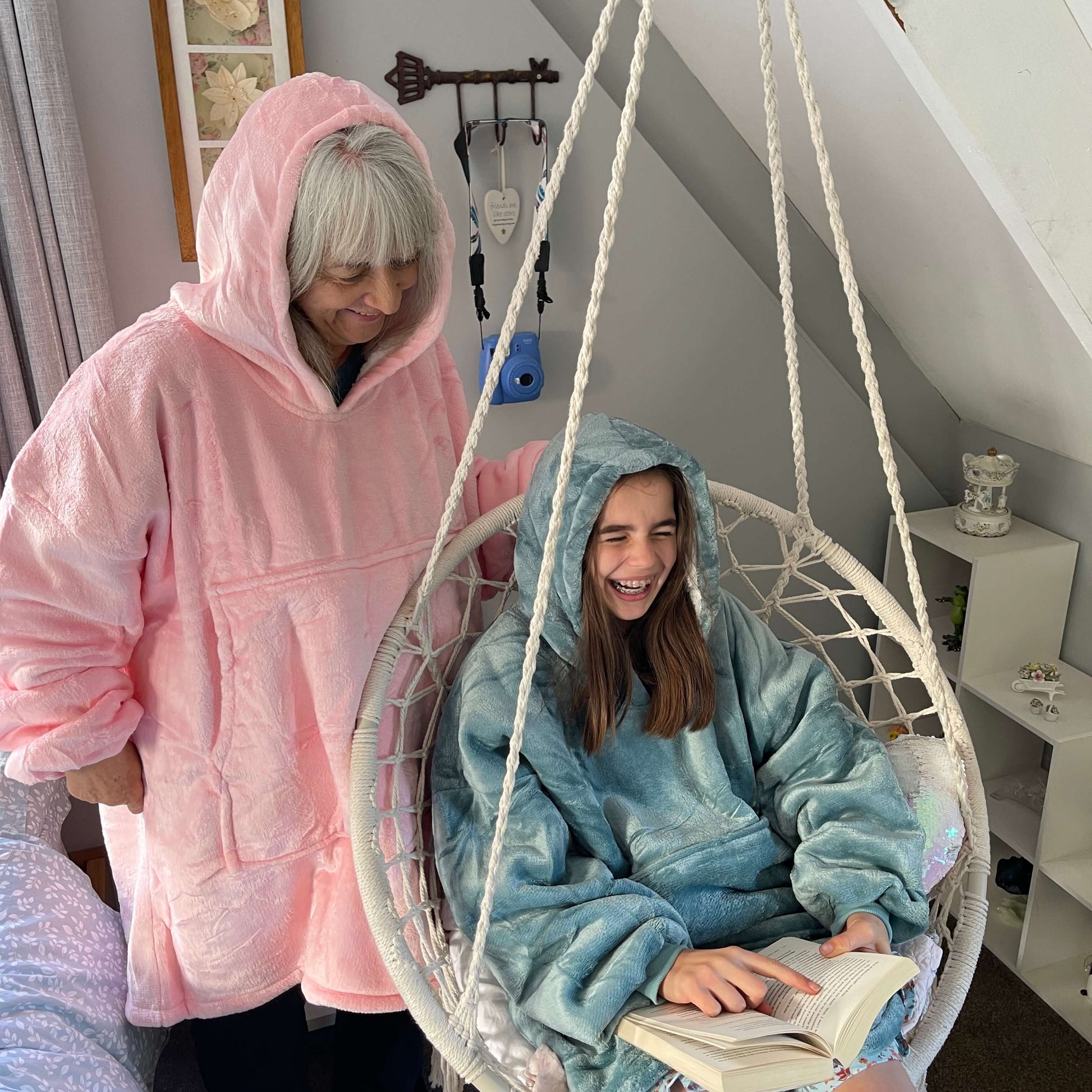 Girl sitting in a swing chair reading a book wearing an oversized blue hoodie. Her grandmother stands behind her wearing an oversized pink hoodie.