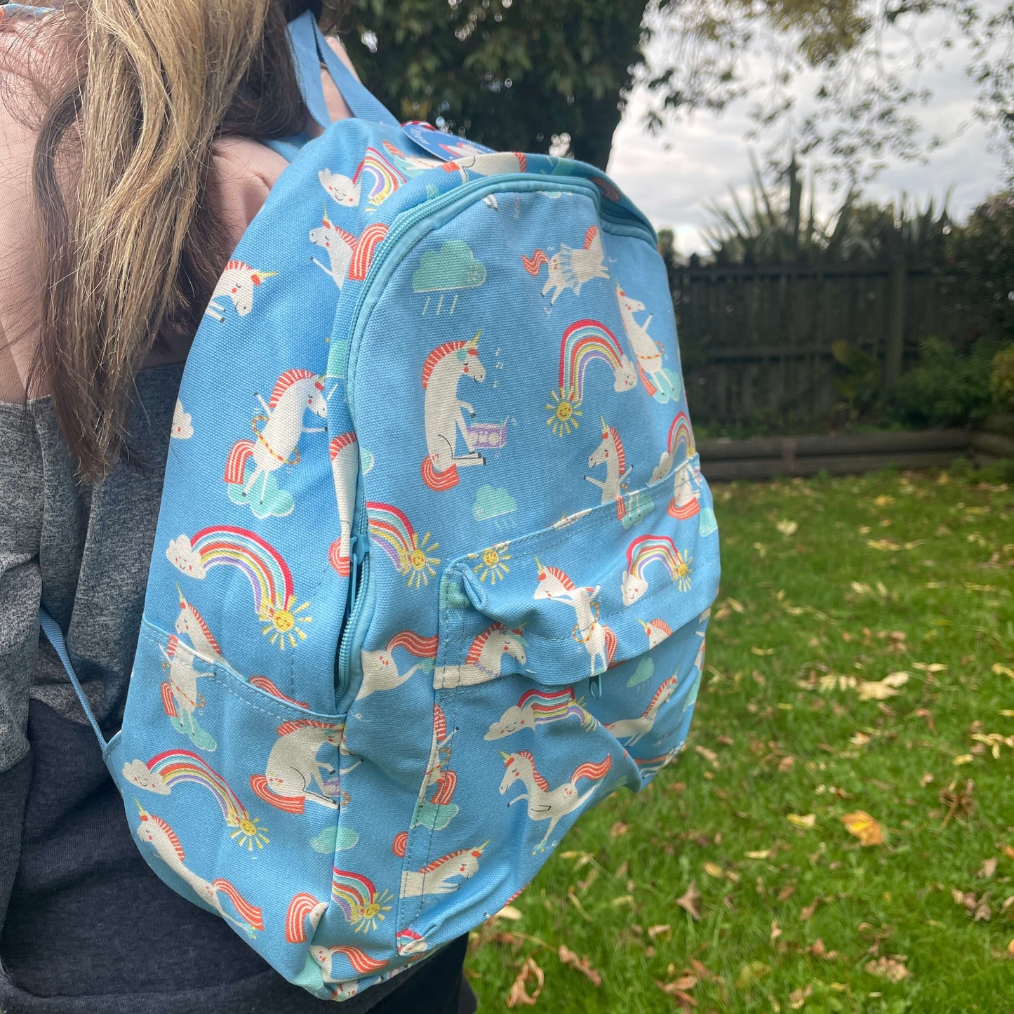 Girl wearing a sky blue backpack with unicorns and rainbows printed on it.