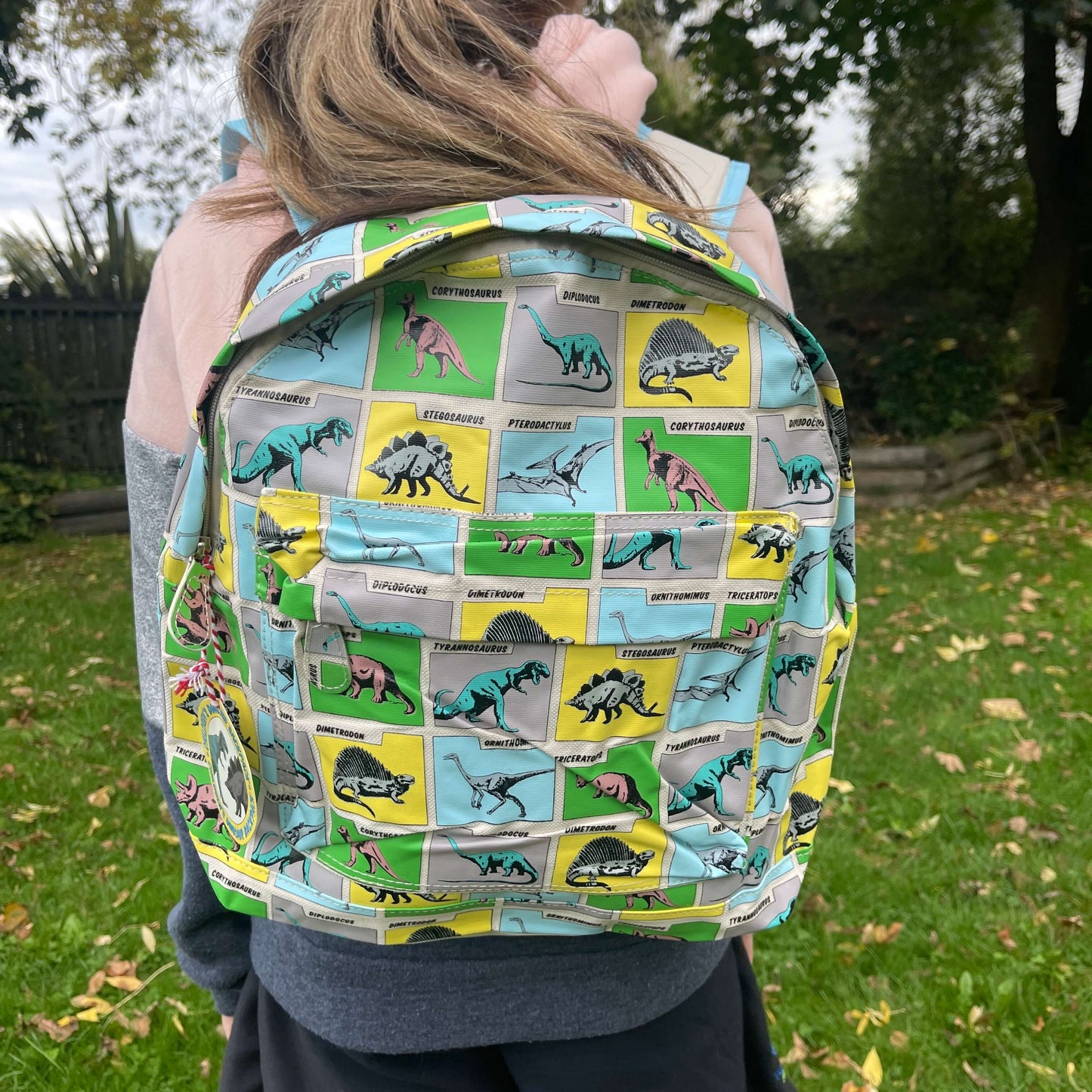 Girl wearing a backpack with dinosaurs printed on it in grey, blue, green & yellow squares.