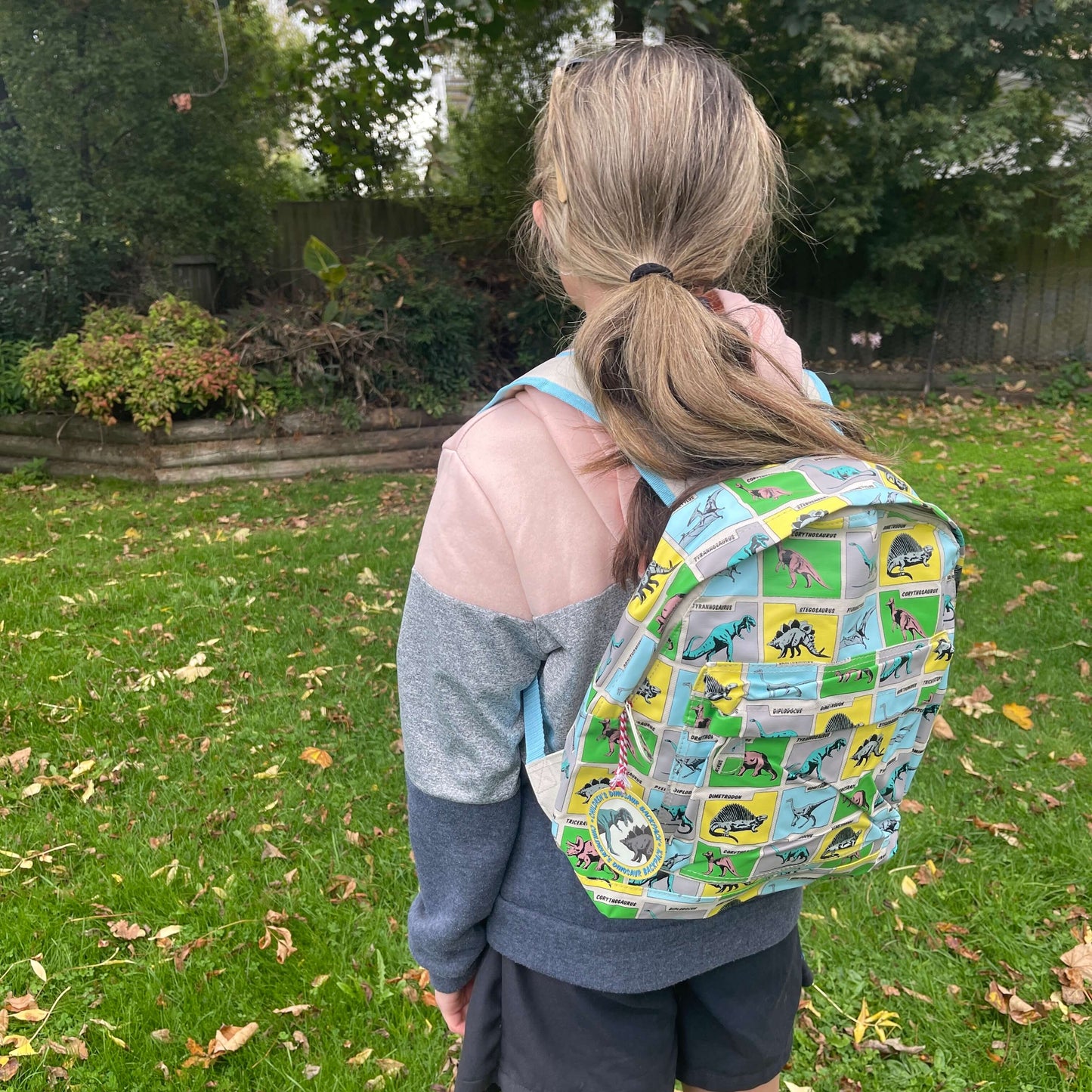 Girl wearing a backpack with dinosaurs printed on it in grey, blue, green & yellow squares.