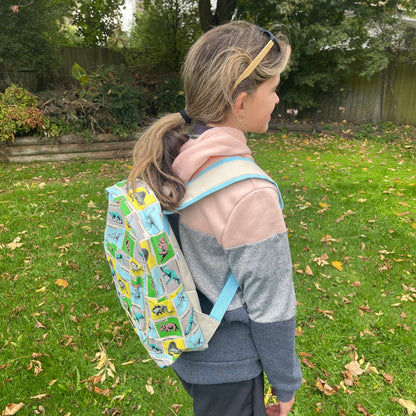 Girl wearing a backpack with dinosaurs printed on it in grey, blue, green & yellow squares.