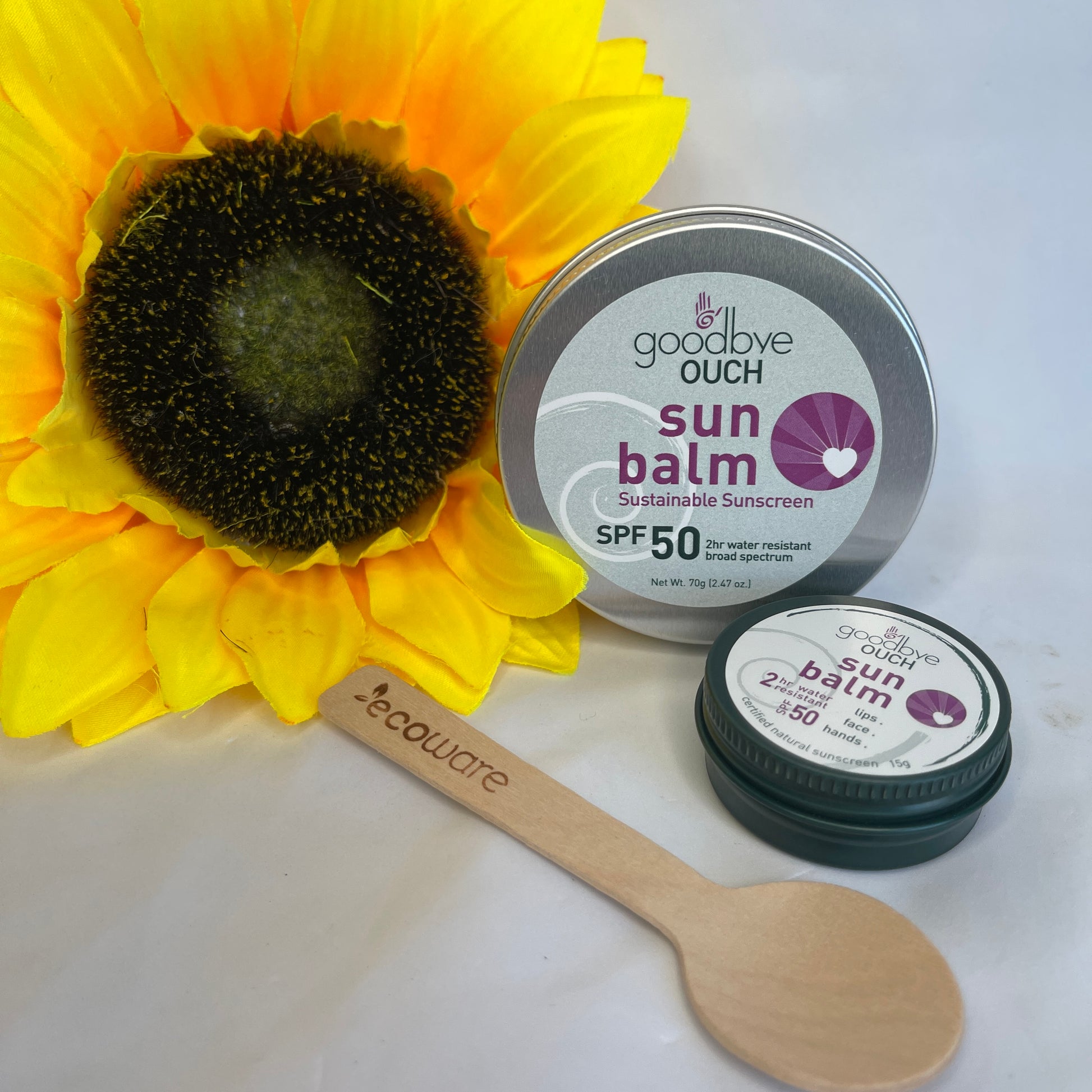 Two tins of sunbalm and a wooden spoon beside a sunflower.