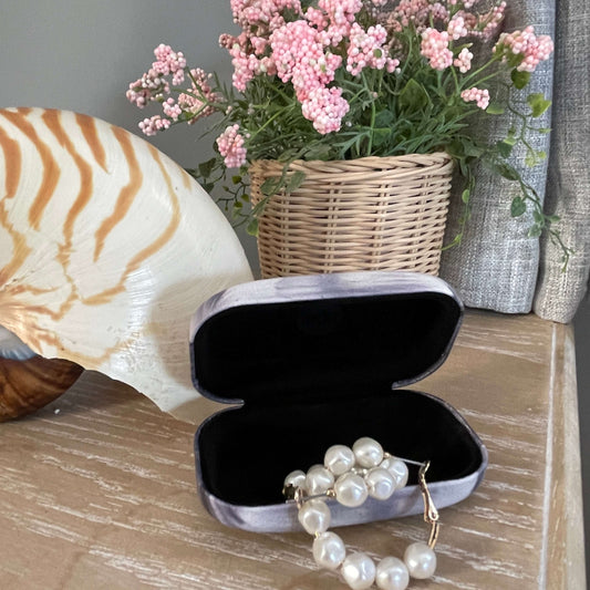 Small trinket case opened with pearl earrings in it sitting on a table with a large shell and faux pot plant.
