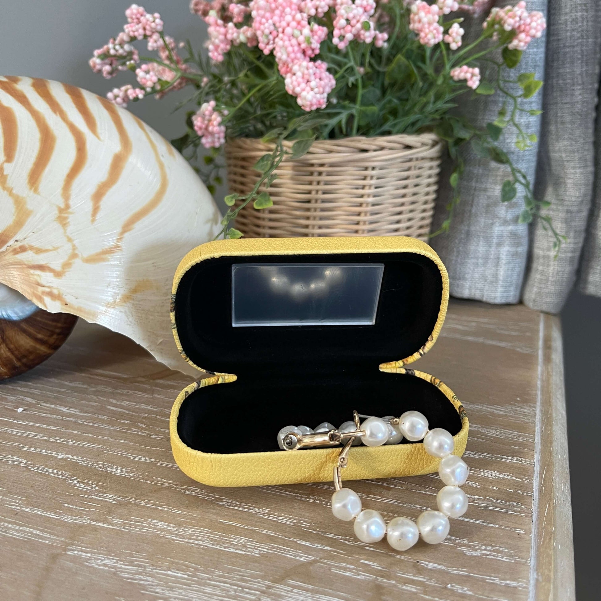 Small trinket case opened showing the mirror and with pearl earrings in it sitting on a table with a large shell and faux pot plant.
