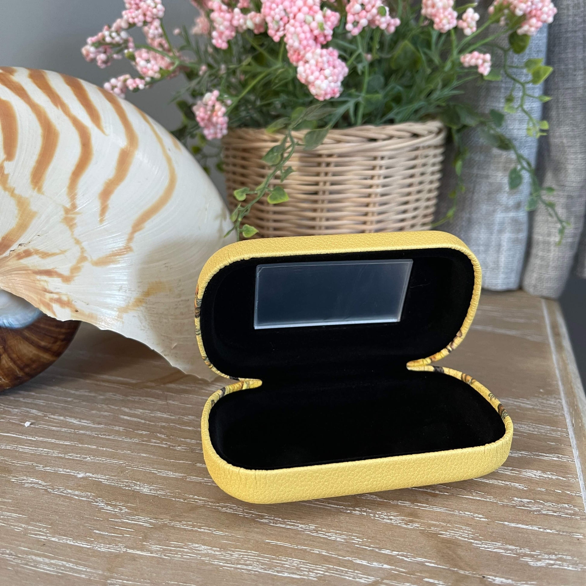 Small trinket case opened showing the mirror and black lining sitting on a table with a large shell and faux pot plant.