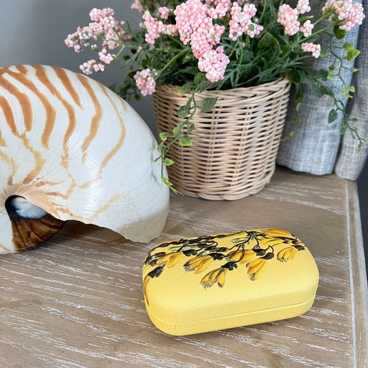 Small trinket case in bright yellow with Kowhai flowers on it sitting on a table with a large shell and faux pot plant.