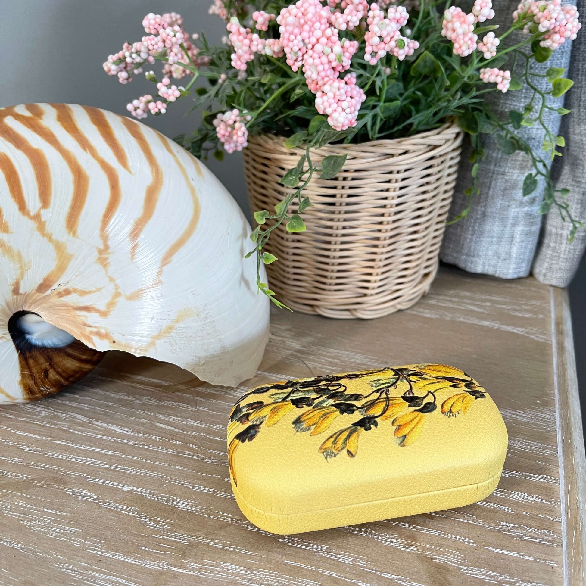 Small trinket case in bright yellow with Kowhai flowers on it sitting on a table with a large shell and faux pot plant.