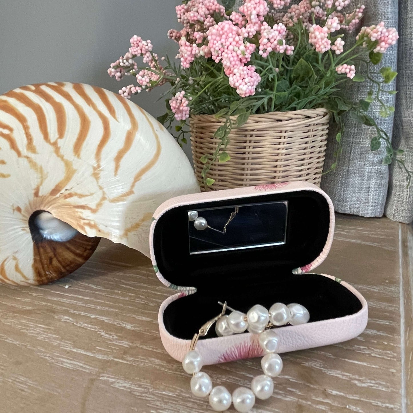 Small trinket case opened showing the mirror and with pearl earrings in it sitting on a table with a large shell and faux pot plant.