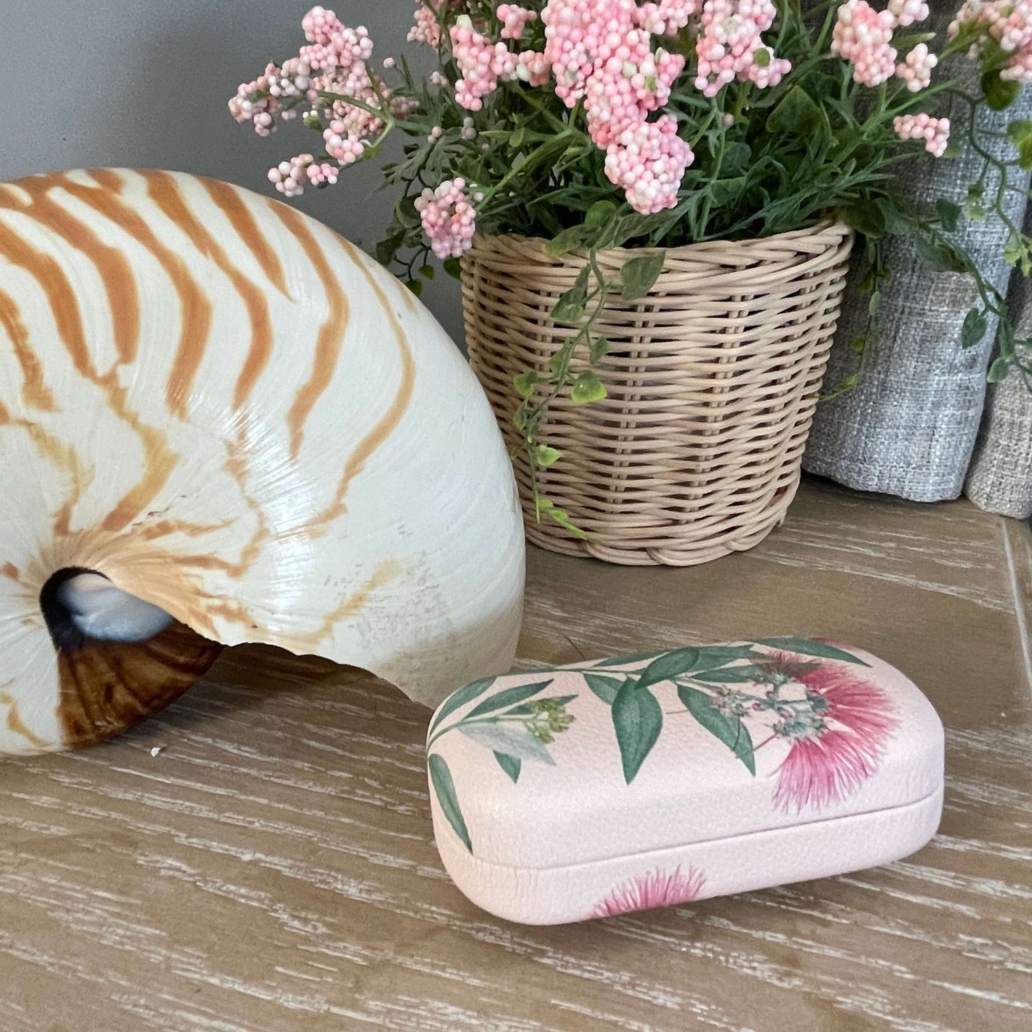 Small trinket case in pale pink with Pohutukawa flowers on it sitting on a table with a large shell and faux pot plant.