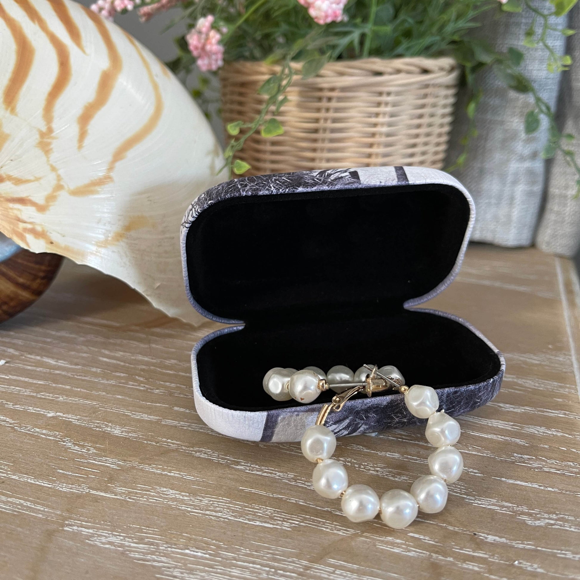 Small trinket case opened with pearl earrings in it sitting on a table with a large shell and faux pot plant.