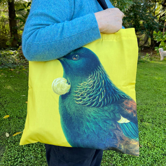 Woman with a tote bag over her arm. The bright yellow tote teatures a Tui bird.