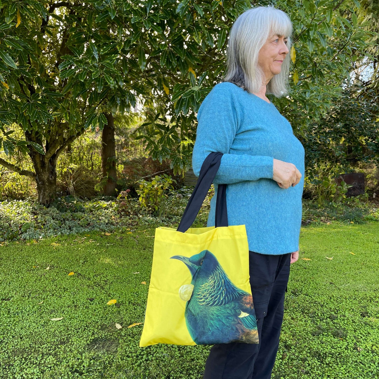 Woman with a tote bag over her arm. The bright yellow tote teatures a Tui bird.