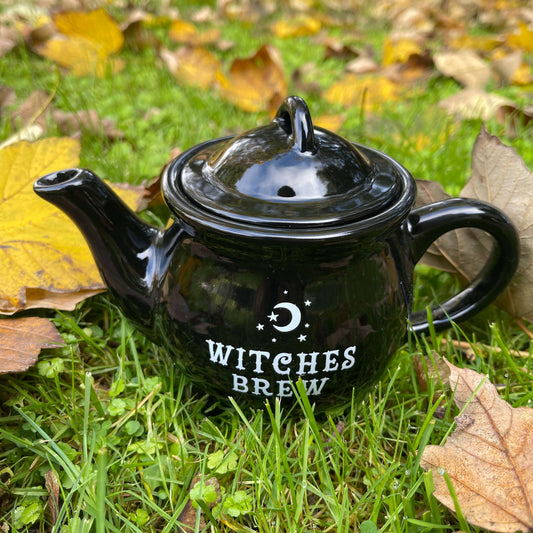Black teapot with Witches Brew written on it in white sitting amongst leaves in the grass.