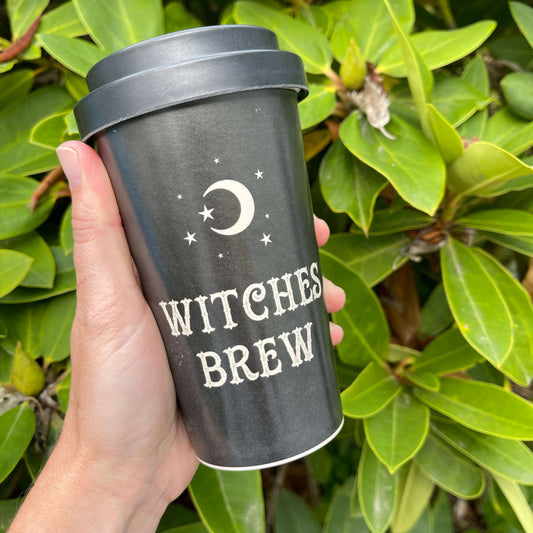 Womens hand holding a Black Bamboo Eco Travel Mug with white font saying "Witches Brew" on it against green foliage.