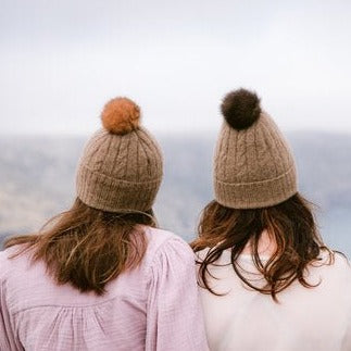 Two people wearing brown knit beanies with pompoms.