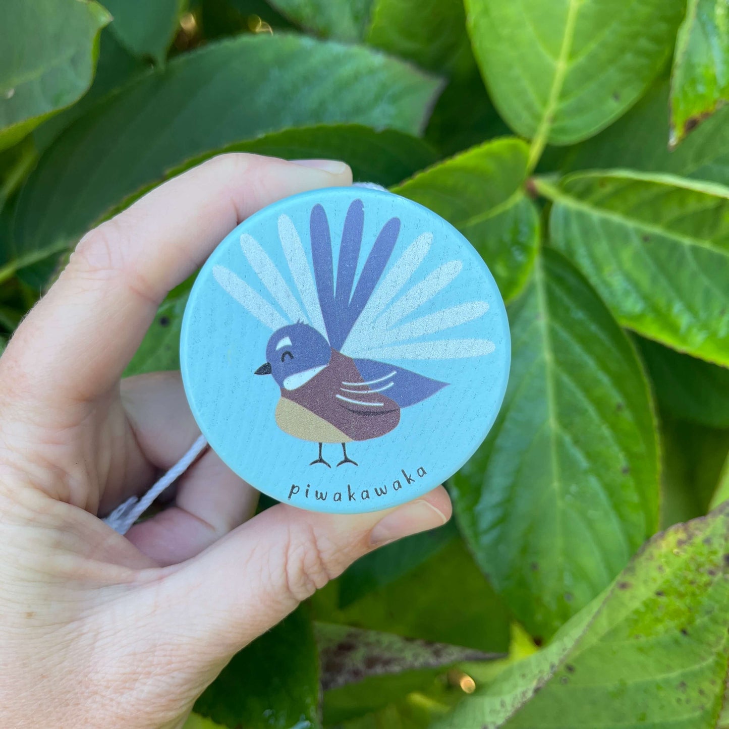 Women's hand holding a sky blue wooden yoyo with a Fantail bird painted on it.
