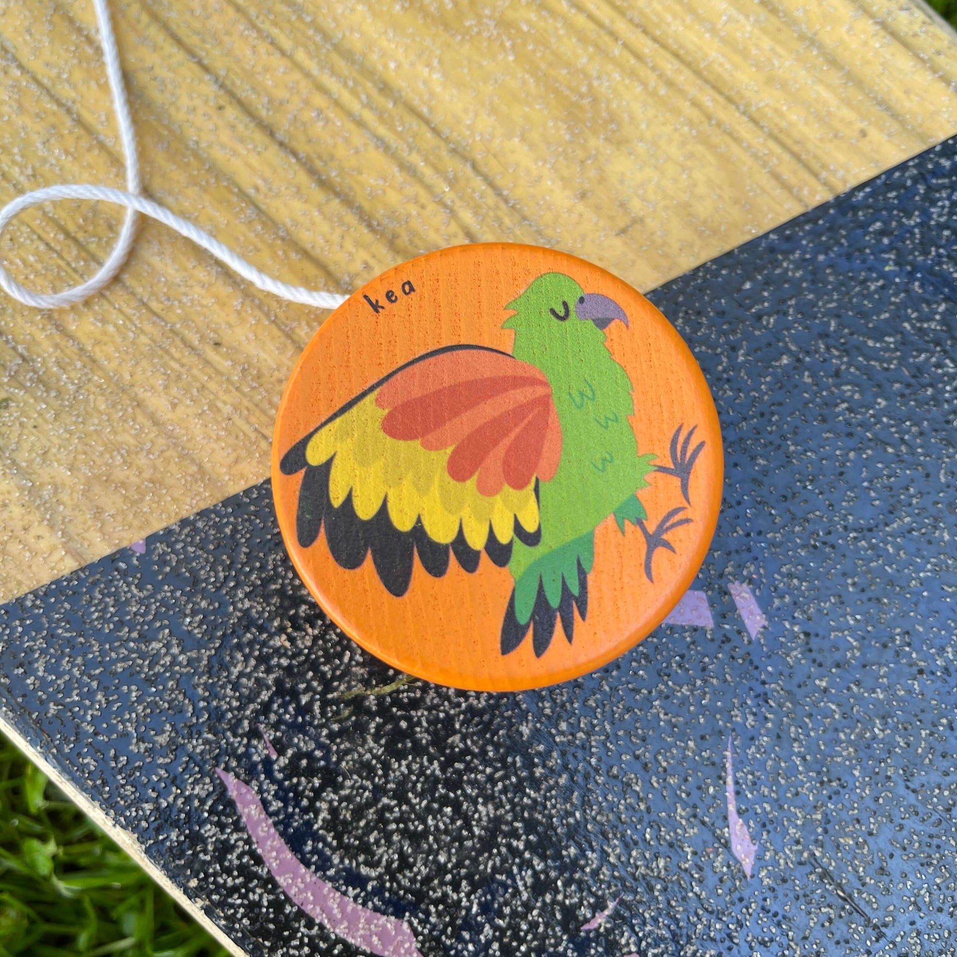 A bright orange wooden yoyo with a Kea bird painted on it sitting on a skateboard.