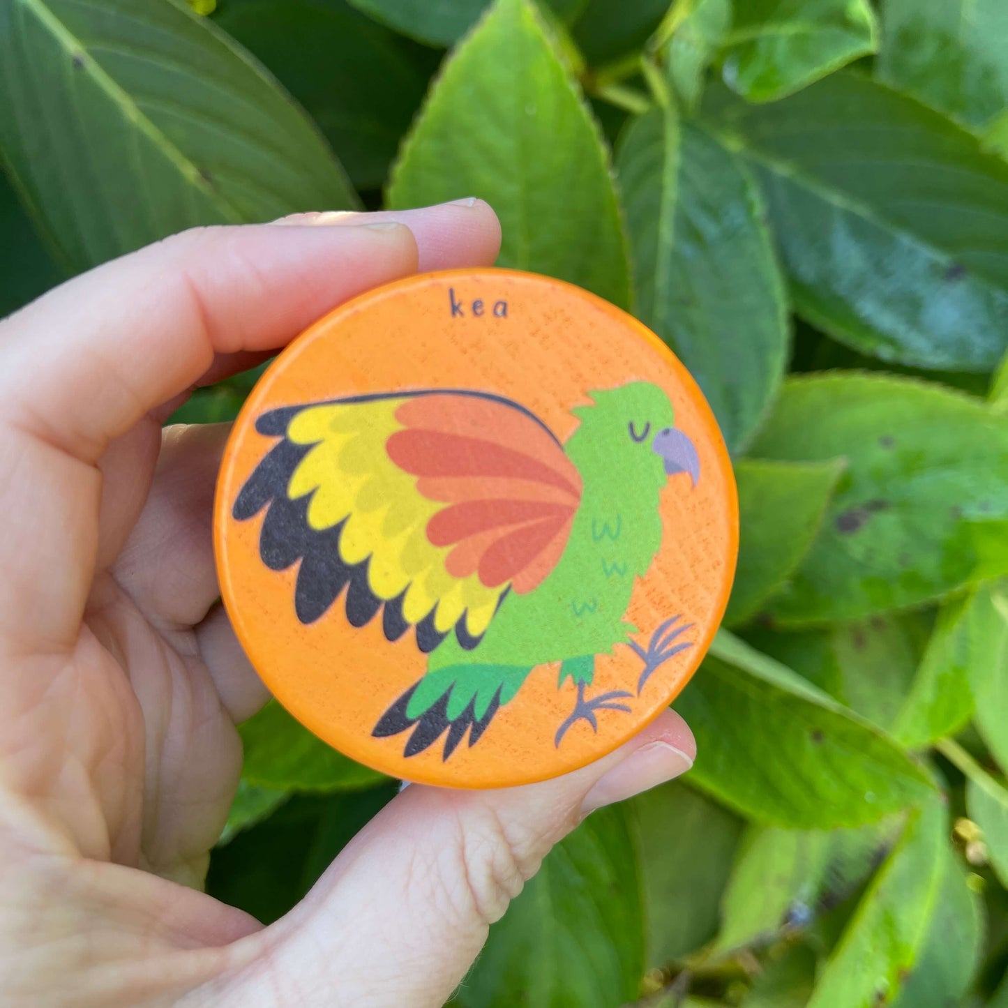 Womens hand holding a bright orange wooden yoyo with a Kea bird painted on it.